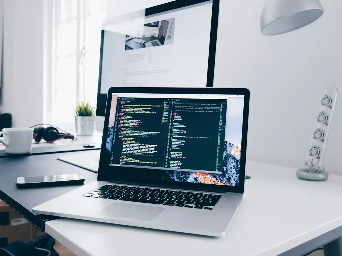 Laptop showing a code editor while a website design is reviewed on a monitor behind it